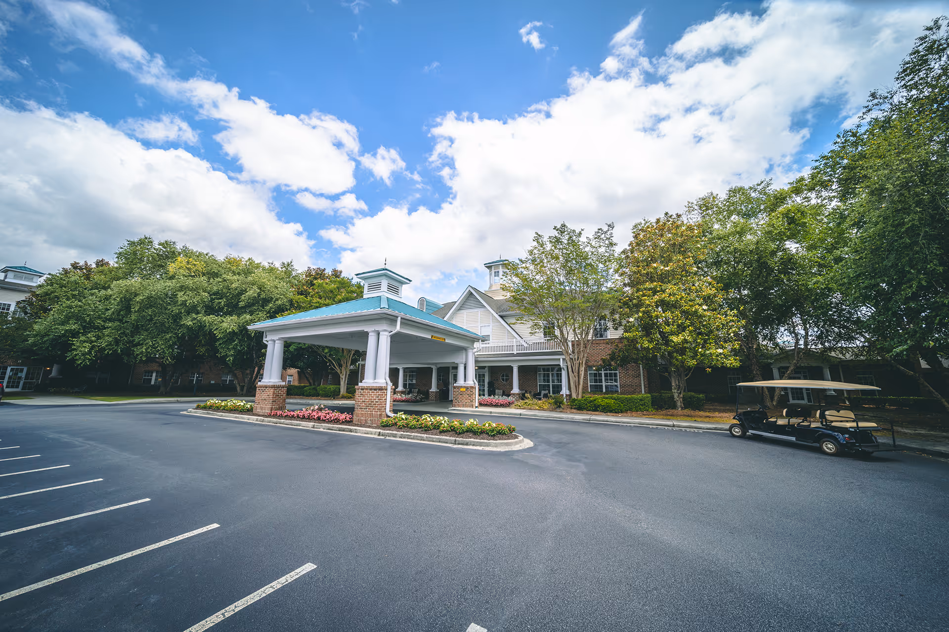 Front entrance of a senior living facility with a covered porte-cochere, landscaped island, parking lot, and a parked golf cart under a partly cloudy sky.