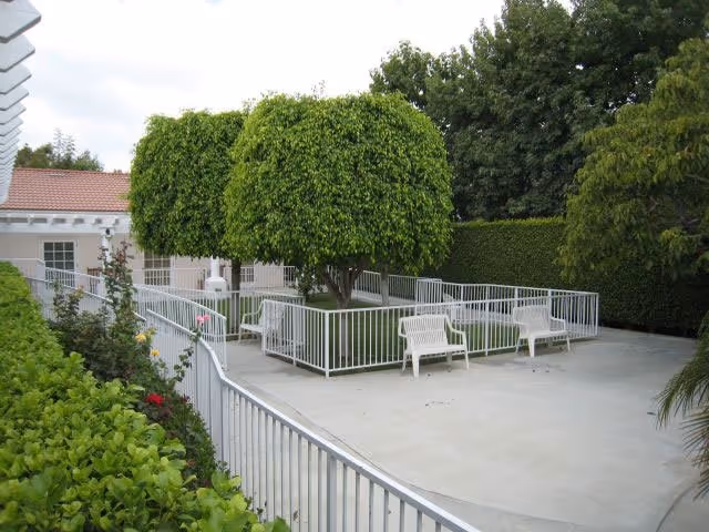 Outdoor patio area with neatly trimmed trees surrounded by white metal railings, two white benches on a concrete surface, and a hedge in the background.