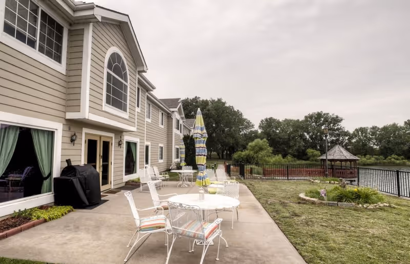 Outdoor patio area at Vintage Park At Waterfront with white metal tables and chairs, some with striped cushions, a closed striped umbrella, a barbecue grill, and a view of a fenced waterfront with trees and a small gazebo in the background.