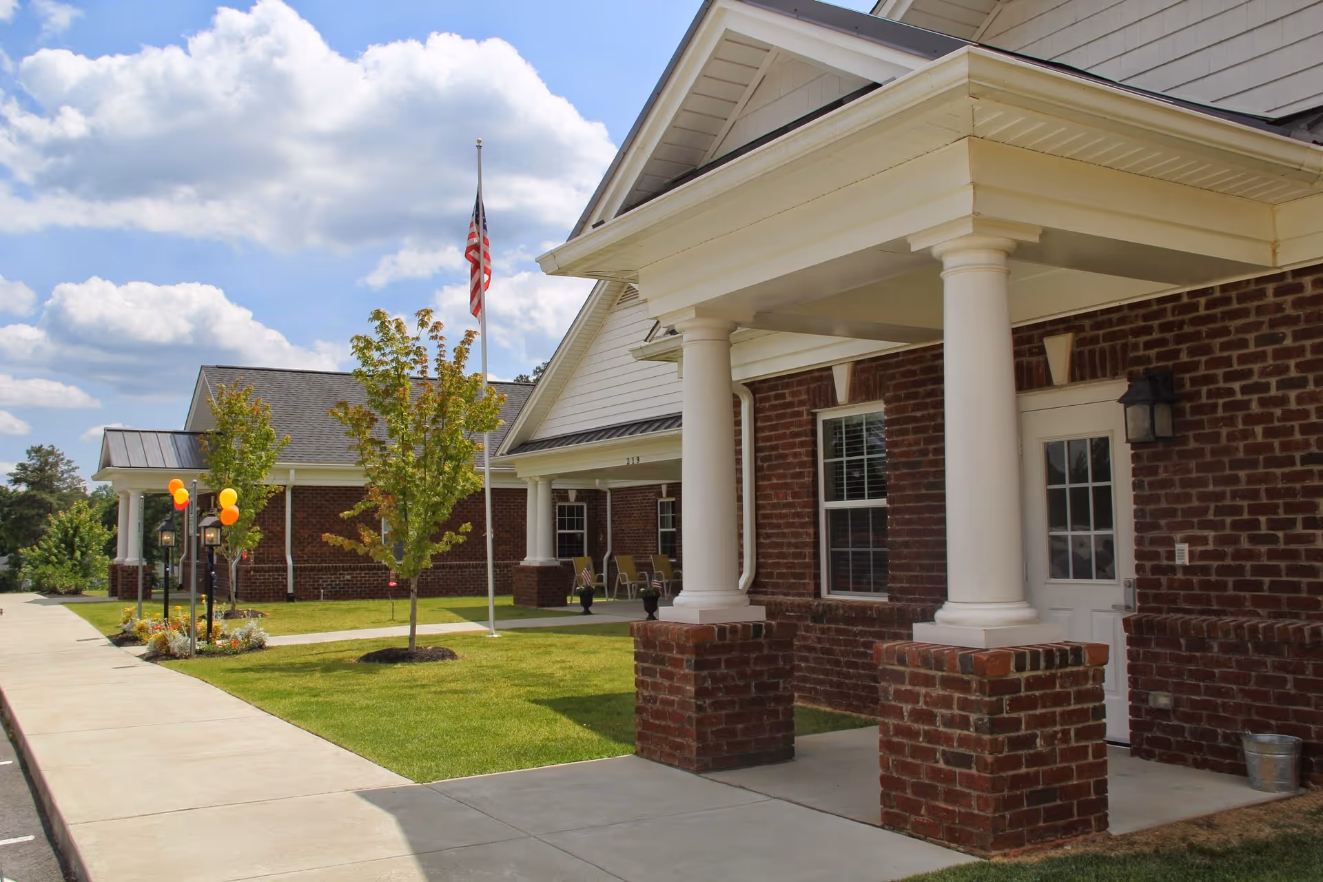Front exterior of a brick senior living building with white columns, an American flag, balloons, and a paved walkway.