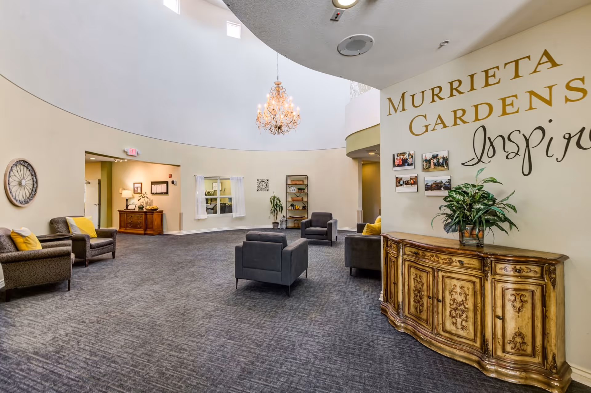 A spacious, well-lit common area in Murrieta Gardens Memory Care featuring a high curved ceiling with a chandelier, several gray armchairs arranged on a carpeted floor, a decorative wooden sideboard with a plant, and wall decor including the facility name and inspirational text.