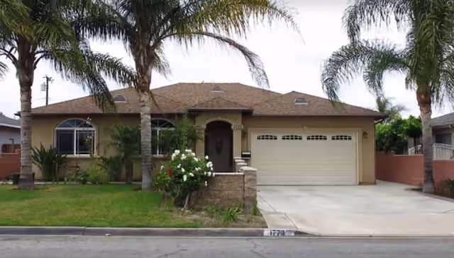 Single-story residential building with a beige exterior, a two-car garage, and a front yard featuring palm trees and green grass.
