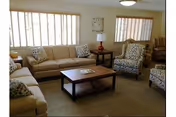 A well-lit living room with beige sofas, patterned armchairs, a wooden coffee table with a magazine on it, and a side table with a lamp. Large windows with vertical blinds allow natural light to fill the room.
