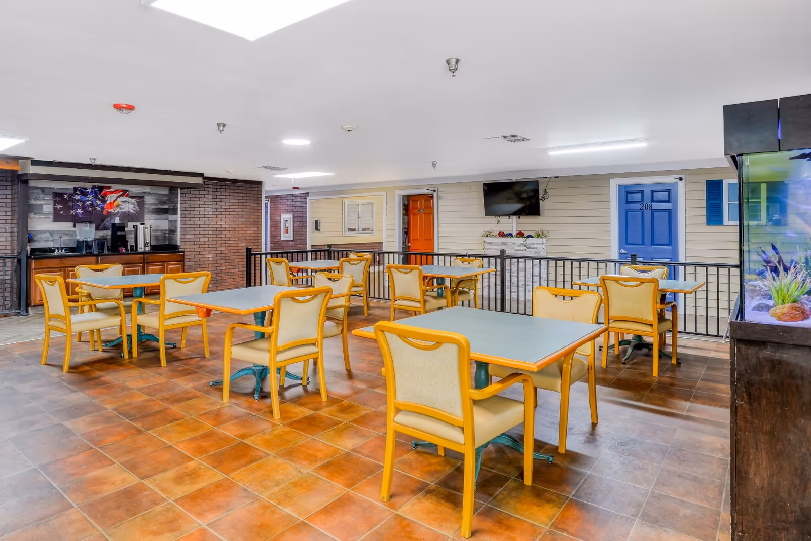 A communal dining area with several tables and yellow cushioned chairs on a tiled floor. The room features a beverage station with an American flag and eagle artwork on the wall, a mounted flat-screen TV, and doors painted in bright colors resembling apartment entrances. There is also a large fish tank visible on the right side.