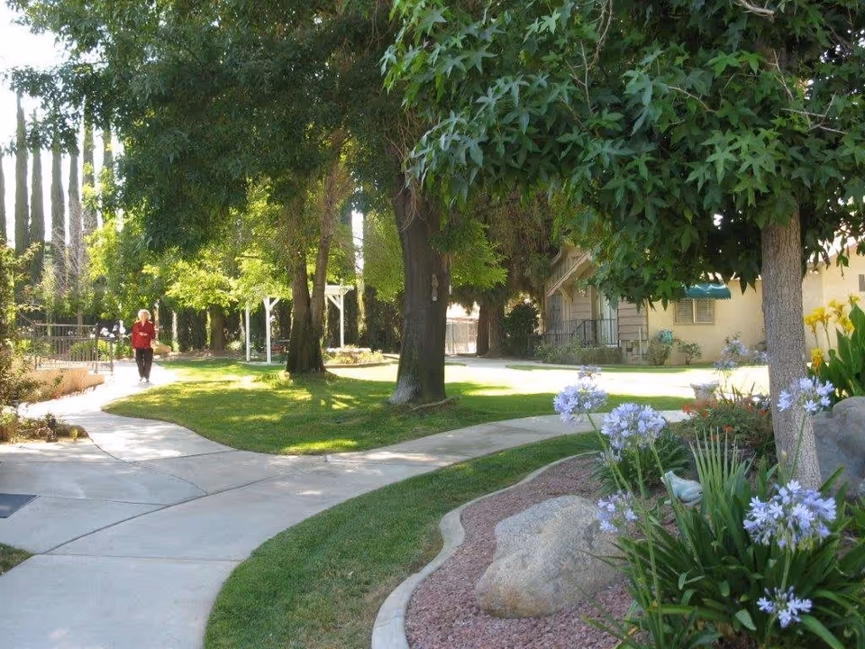 A sunny outdoor garden area with a winding concrete pathway surrounded by green grass, trees, and flowering plants. A person in a red jacket is walking along the path. There are buildings visible in the background.