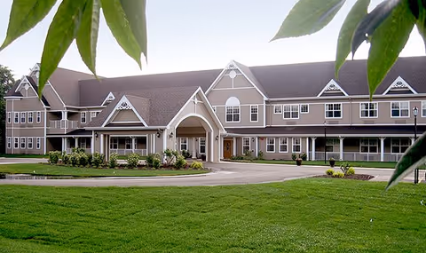 Front exterior view of a large, two-story senior living facility building with a peaked roof, multiple windows, and a covered entrance. The building is surrounded by a well-maintained lawn and some landscaping, with tree leaves partially framing the top of the image.