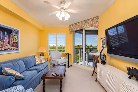 A cozy living room with yellow walls featuring a blue couch with floral pillows, a wooden coffee table, a white armchair, a ceiling fan with lights, a large flat-screen TV mounted on the wall, and a sliding glass door leading to a balcony with outdoor seating and a view of greenery.