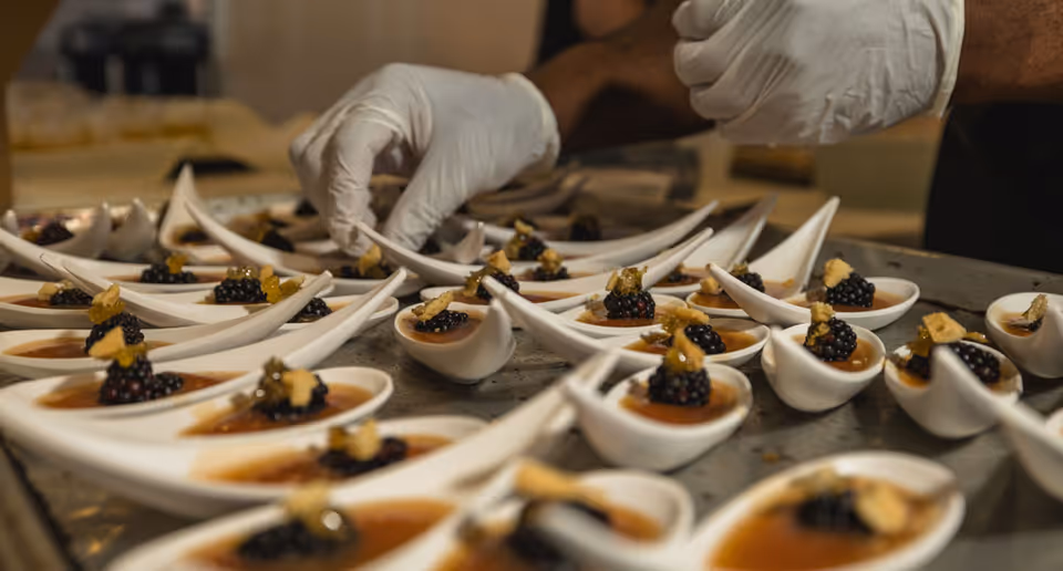 Close-up of a person wearing white gloves carefully placing garnishes on small white ceramic spoons filled with a dessert topped with blackberries and a small crunchy garnish, arranged on a metal tray.