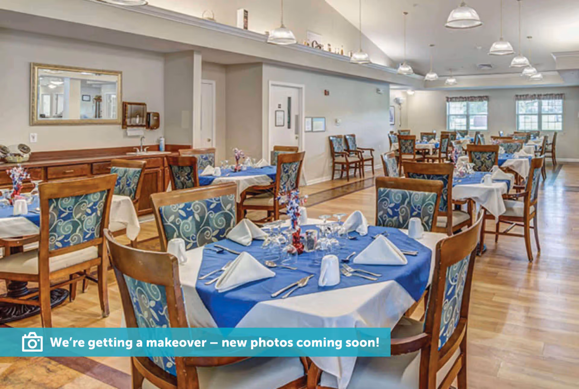 Spacious dining room with multiple tables set with blue tablecloths, napkins, wooden chairs, and overhead pendant lights.