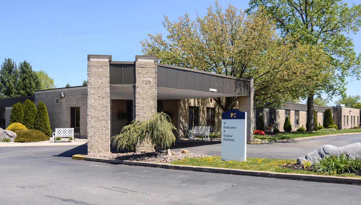 Front entrance of a single-story brick nursing facility with a covered drop-off, sign, benches, and landscaped grounds.
