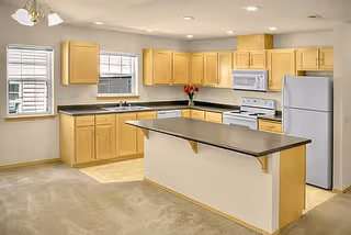 Bright open kitchen with light wood cabinets, a central island, white appliances, and two windows.