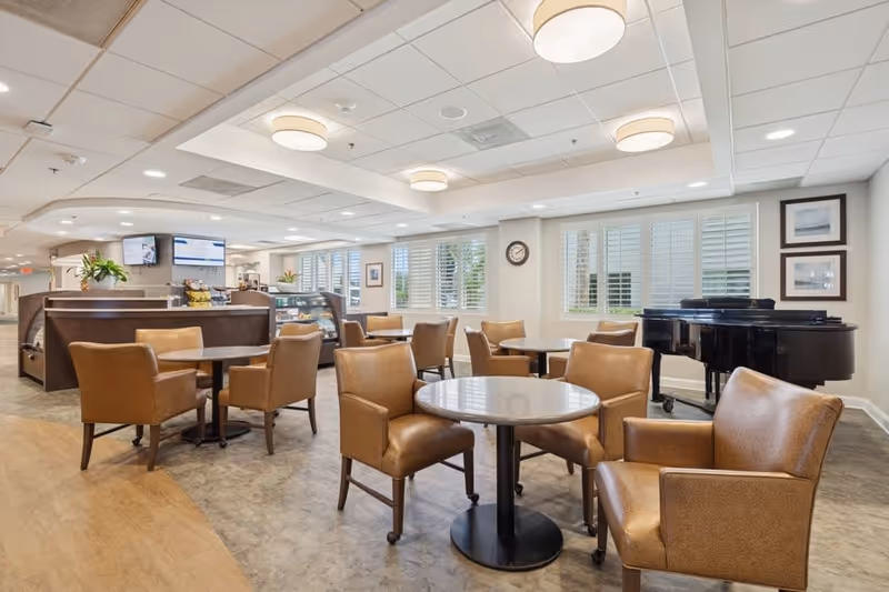 A bright and spacious common area with several round tables and brown leather chairs arranged around them. In the background, there is a counter with a display case and a few plants. Large windows with white shutters let in natural light. A black grand piano is positioned near the windows, and framed pictures hang on the wall.