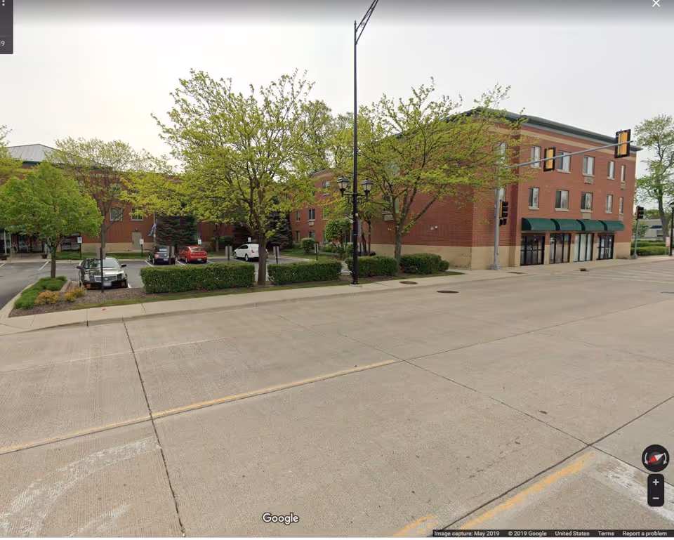 Three-story brick apartment building with green awnings, trees, and a small parking lot beside a wide street.