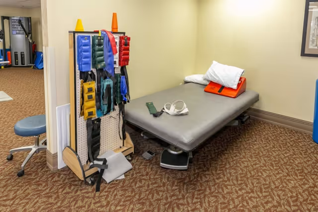 Small therapy room with a padded treatment table, a rack of colorful rehab bands and weights, and a rolling stool.