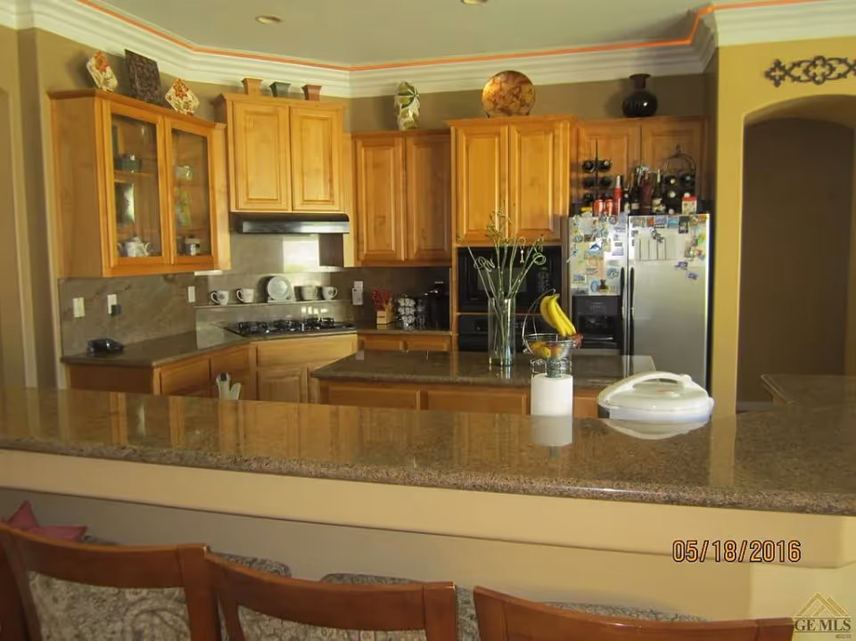 Kitchen with wooden cabinets, a large granite countertop island, stainless refrigerator, and bar stools.