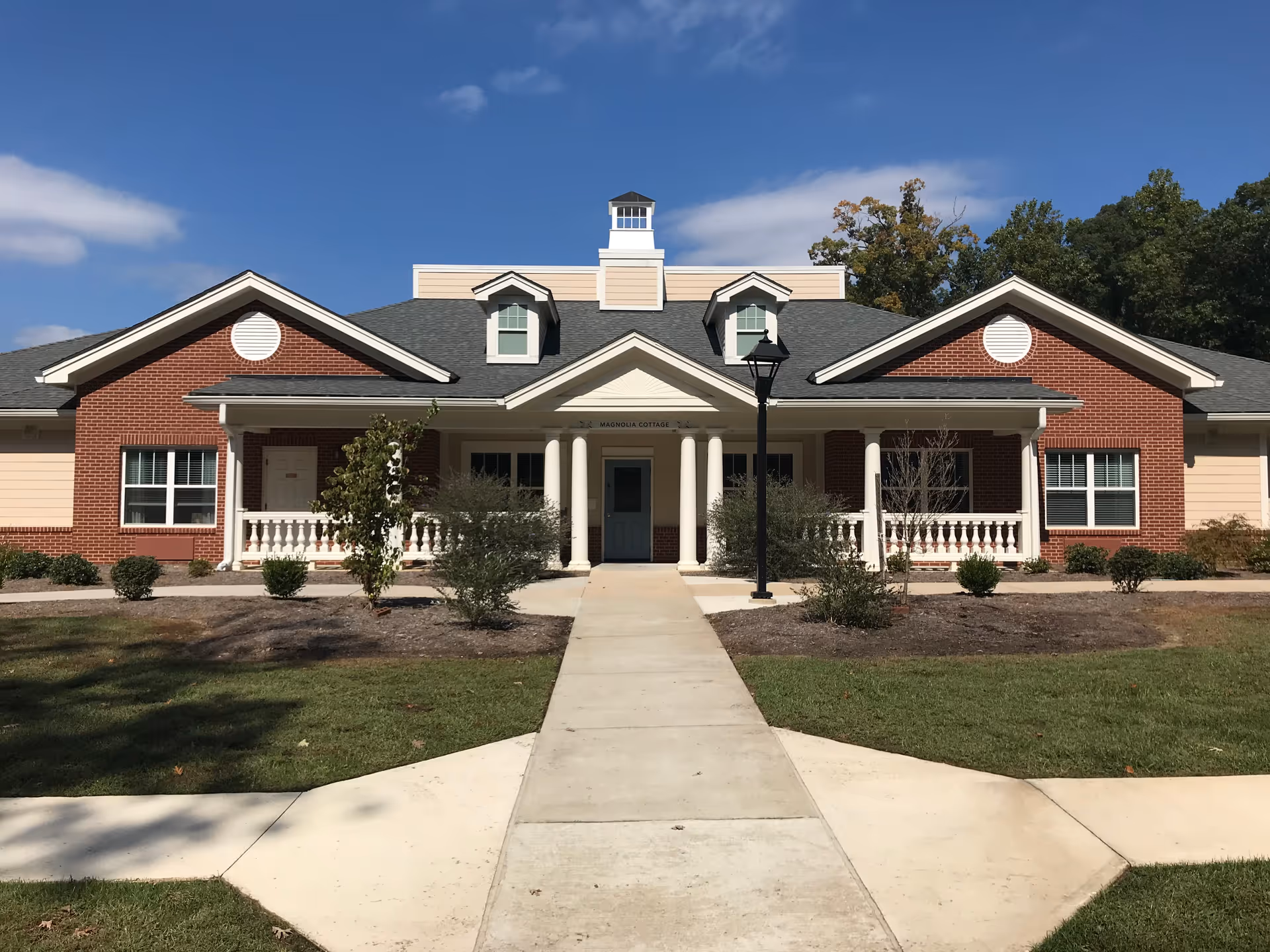 Front exterior view of a single-story brick and siding building with a central entrance featuring white columns and a small cupola on the roof. There is a concrete walkway leading up to the entrance, surrounded by grass and small shrubs under a clear blue sky.