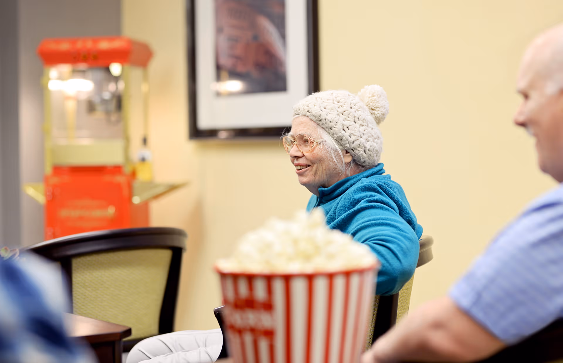 An elderly woman wearing a white knitted hat and blue jacket is sitting and smiling in a room with a popcorn machine and a large bucket of popcorn in the foreground. Another elderly person is partially visible sitting nearby.