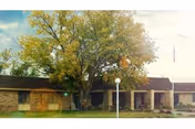Exterior view of a single-story brick building with a large tree in front, a grassy lawn, and a white lamppost near the entrance under a partly cloudy sky.