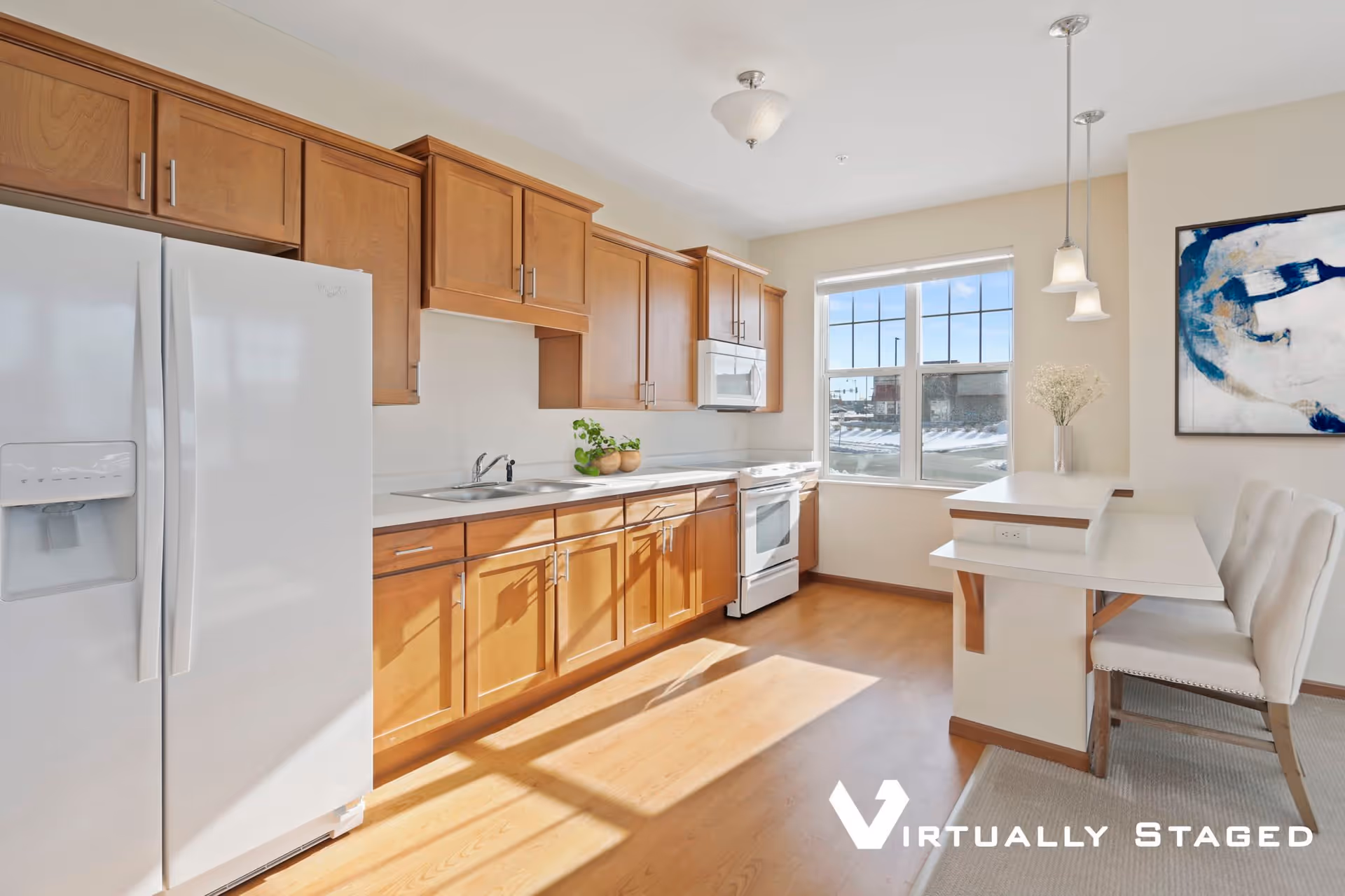 Bright kitchen with wooden cabinets, white appliances including a refrigerator, stove, and microwave. There is a window letting in natural light, a small breakfast bar with two white chairs, and a piece of abstract blue and white artwork on the wall.