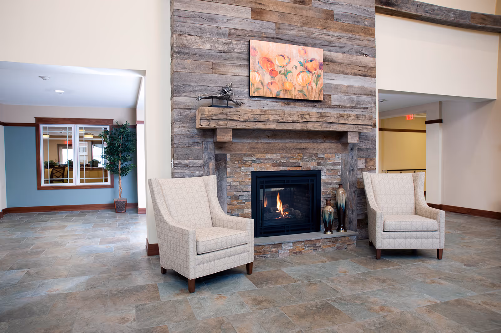 A cozy seating area with two beige patterned armchairs facing a stone fireplace with a wooden mantel. Above the mantel is a colorful painting of flowers. The floor is tiled, and there is a potted plant visible in the background through an open doorway.