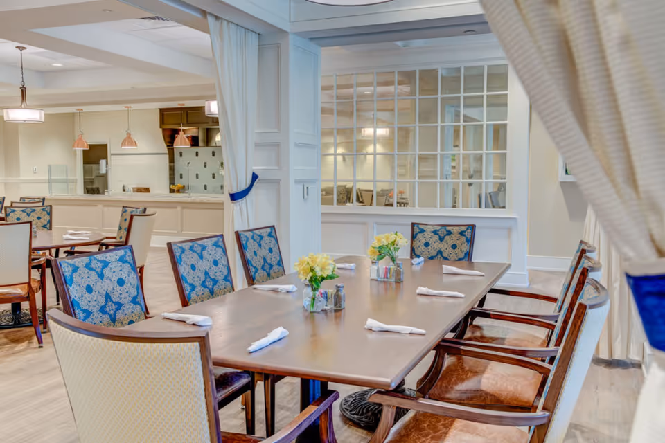 A well-lit dining room with a long wooden table set with napkins and small flower arrangements. The chairs around the table have patterned blue and beige upholstery. In the background, there is a kitchen area with hanging pendant lights and additional tables and chairs. The room features light-colored walls and a large window with multiple panes separating the dining area from another room.