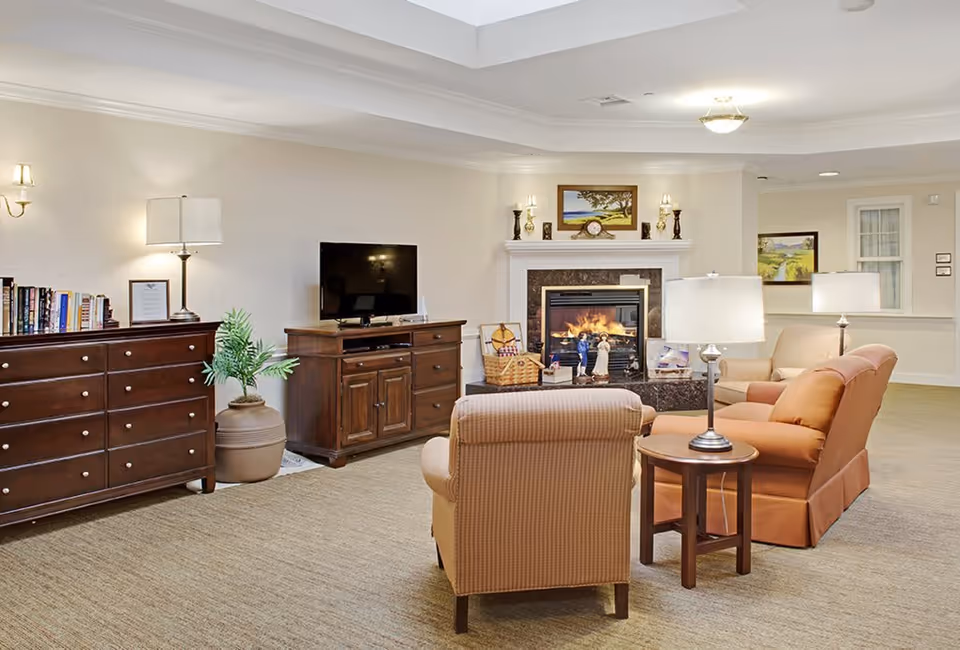 A cozy living room area with a beige armchair and an orange sofa facing a fireplace with a fire burning. There is a wooden TV stand with a flat-screen TV, a wooden dresser with books and a lamp, and a potted plant. The room is softly lit with table lamps and wall sconces, and there are framed paintings on the walls.