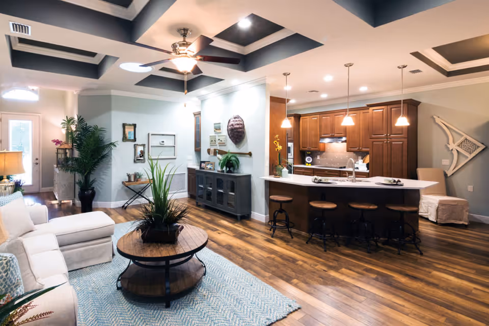 Open concept living area with a white sectional sofa, a round wooden coffee table with a plant centerpiece, and a kitchen with wooden cabinets and a white countertop island with four bar stools. The ceiling features a tray design with recessed lighting and a ceiling fan. The room has wooden flooring and light-colored walls with decorative wall art and plants.