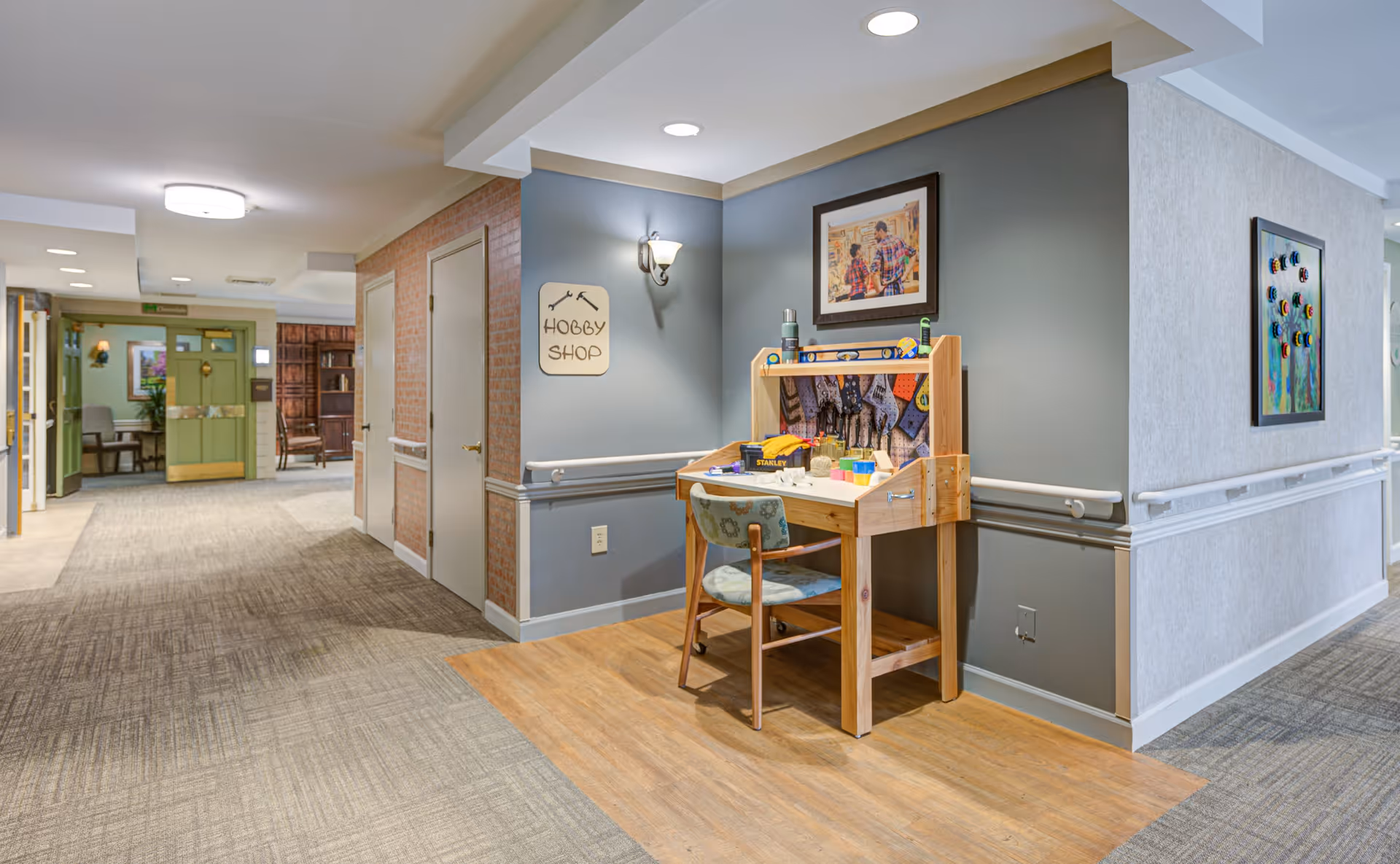A hallway in a senior living facility with a small wooden hobby shop workstation and chair against a gray wall. The wall has a sign labeled 'Hobby Shop' and a framed picture above the workstation. The hallway has carpeted floors with a wooden section under the workstation, handrails along the walls, and doors leading to other rooms. The far end of the hallway shows an open area with more seating and decor.