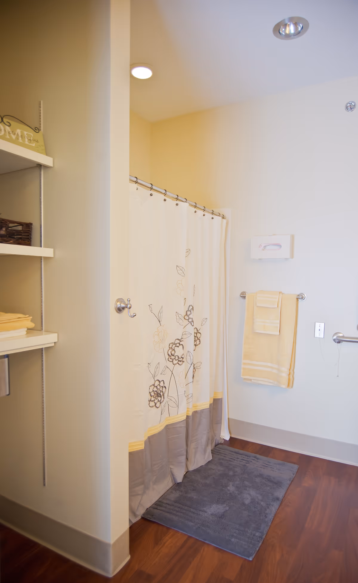 A bathroom with a shower area enclosed by a floral-patterned shower curtain. There is a gray bath mat on the wooden floor in front of the shower. On the right wall, a towel rack holds two yellow towels, and a tissue dispenser is mounted above it. To the left, there are shelves with folded towels and a decorative sign.