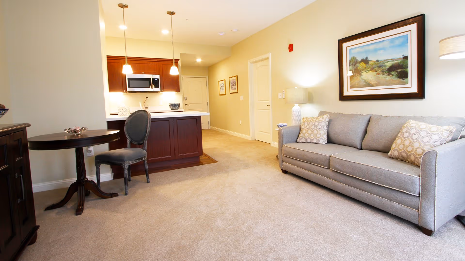 A cozy senior living space featuring a gray sofa with patterned cushions, a floor lamp, and a framed landscape painting on the wall. To the left, there is a small round wooden table with a single upholstered chair. In the background, a kitchen area with wooden cabinets, a microwave, and pendant lights is visible.