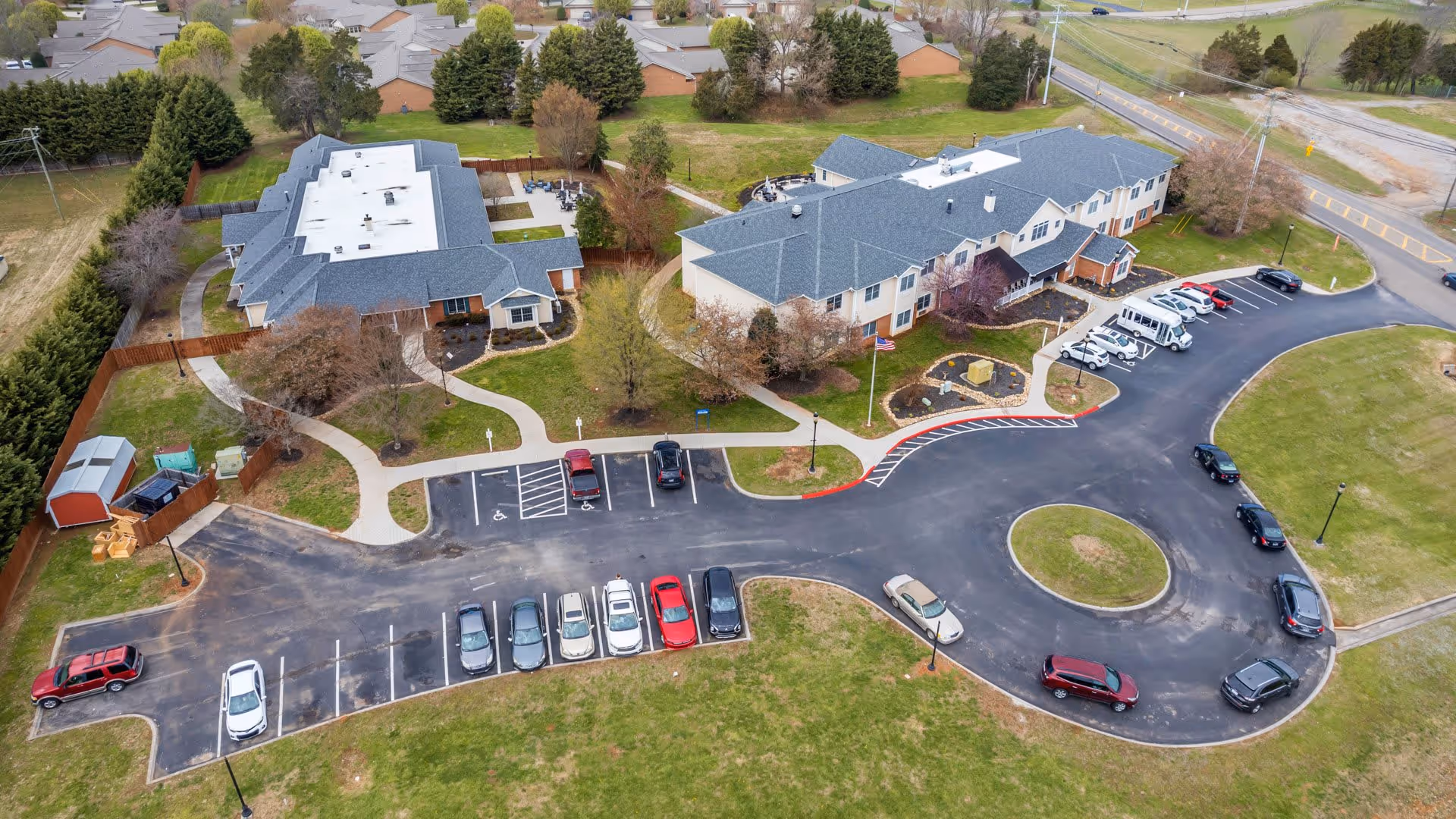 Aerial view of Wellington Manor Knoxville showing two large buildings with gray roofs surrounded by trees and pathways. There is a parking lot with several cars parked and a circular driveway in front of the buildings. The area is bordered by grass and residential houses are visible in the background.