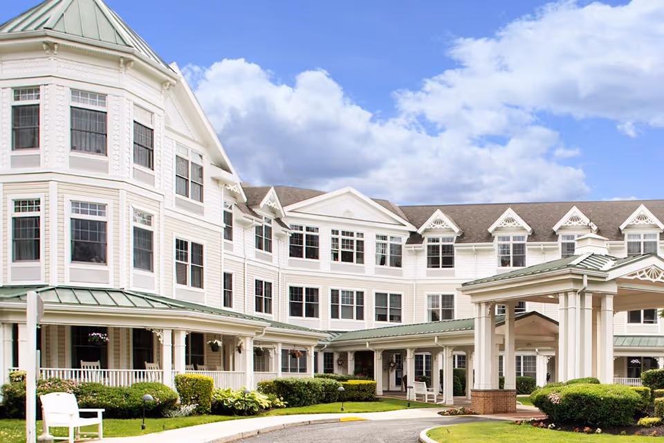 White multi-story senior living building with a covered porte-cochère entrance, wraparound porch and landscaped grounds under a blue sky.