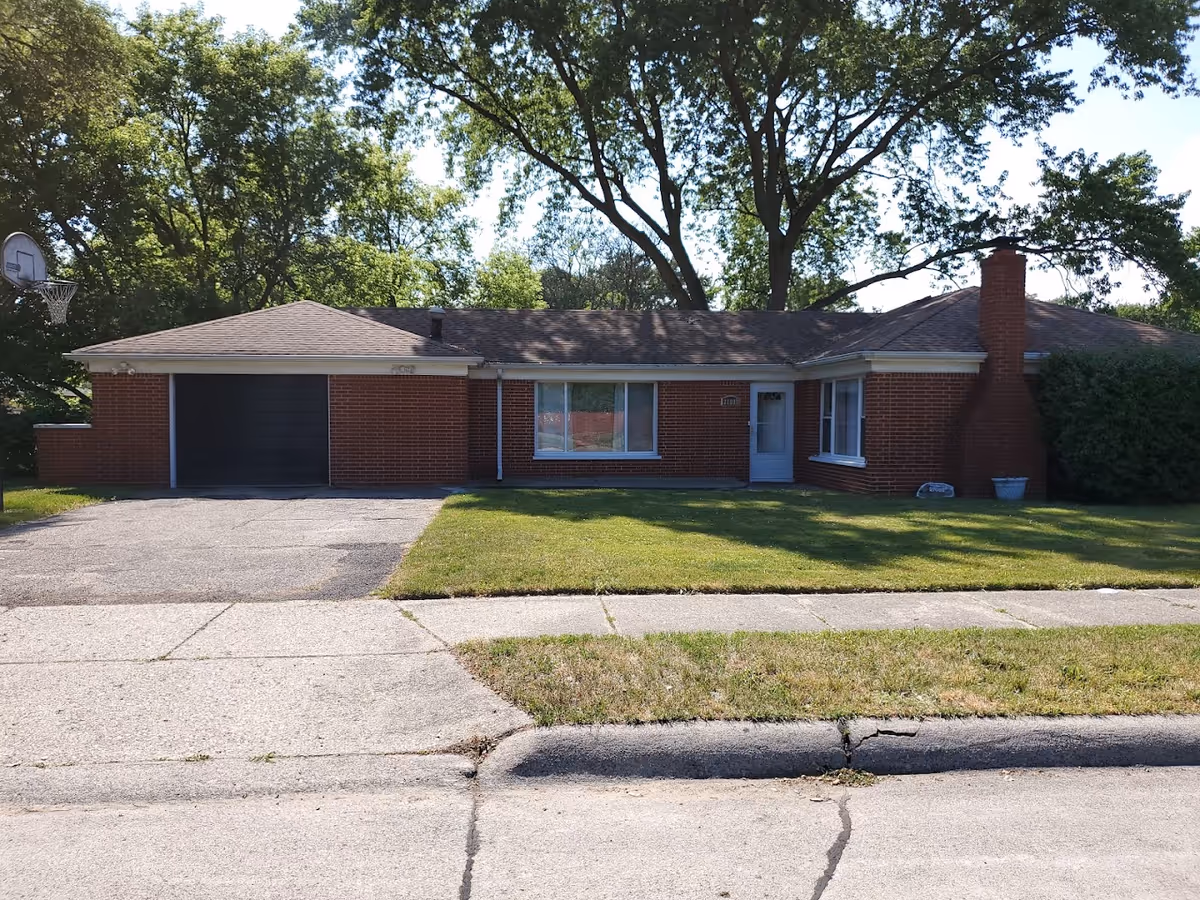 Single-story brick ranch house front with attached garage, driveway, and a grassy lawn under large trees.