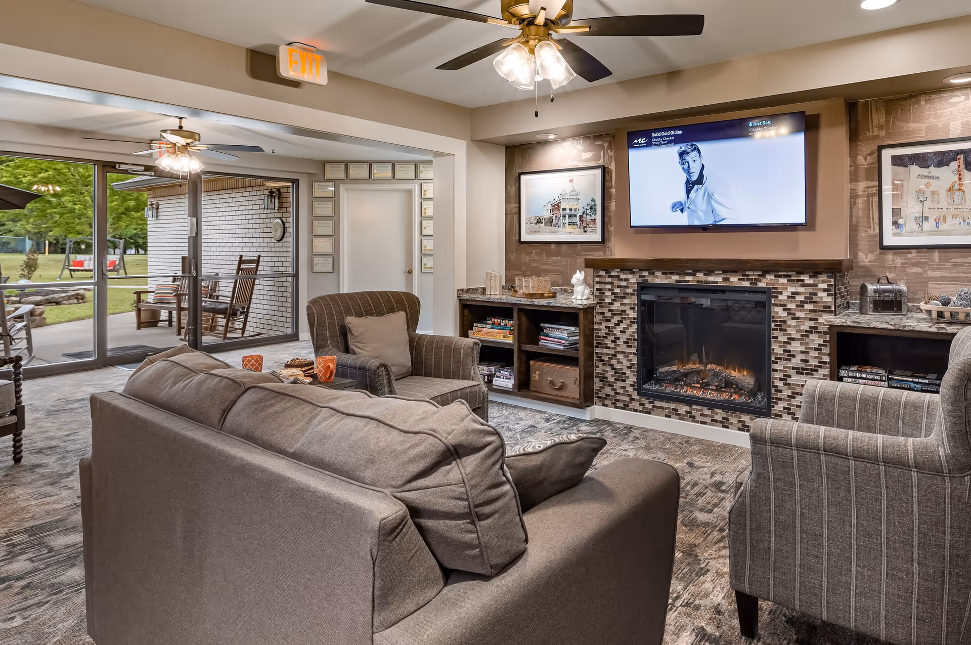A cozy living room area with a gray sofa and two armchairs arranged around a modern electric fireplace with a mosaic tile surround. Above the fireplace is a flat-screen TV displaying a black and white image of a man. The room has a ceiling fan with lights, framed artwork on the walls, and built-in shelves with books and decorative items. Large sliding glass doors open to an outdoor patio with rocking chairs and a view of green trees and grass.