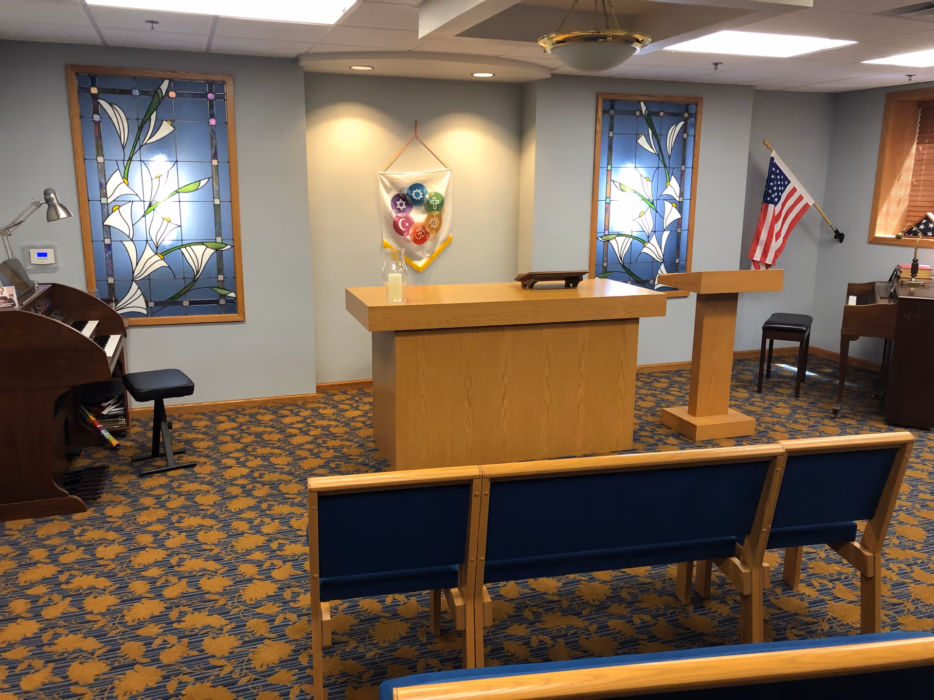 Interior of a small chapel or meditation room with wooden pews, a wooden altar and lectern, stained glass windows with white lilies, an American flag, and a piano with a stool.