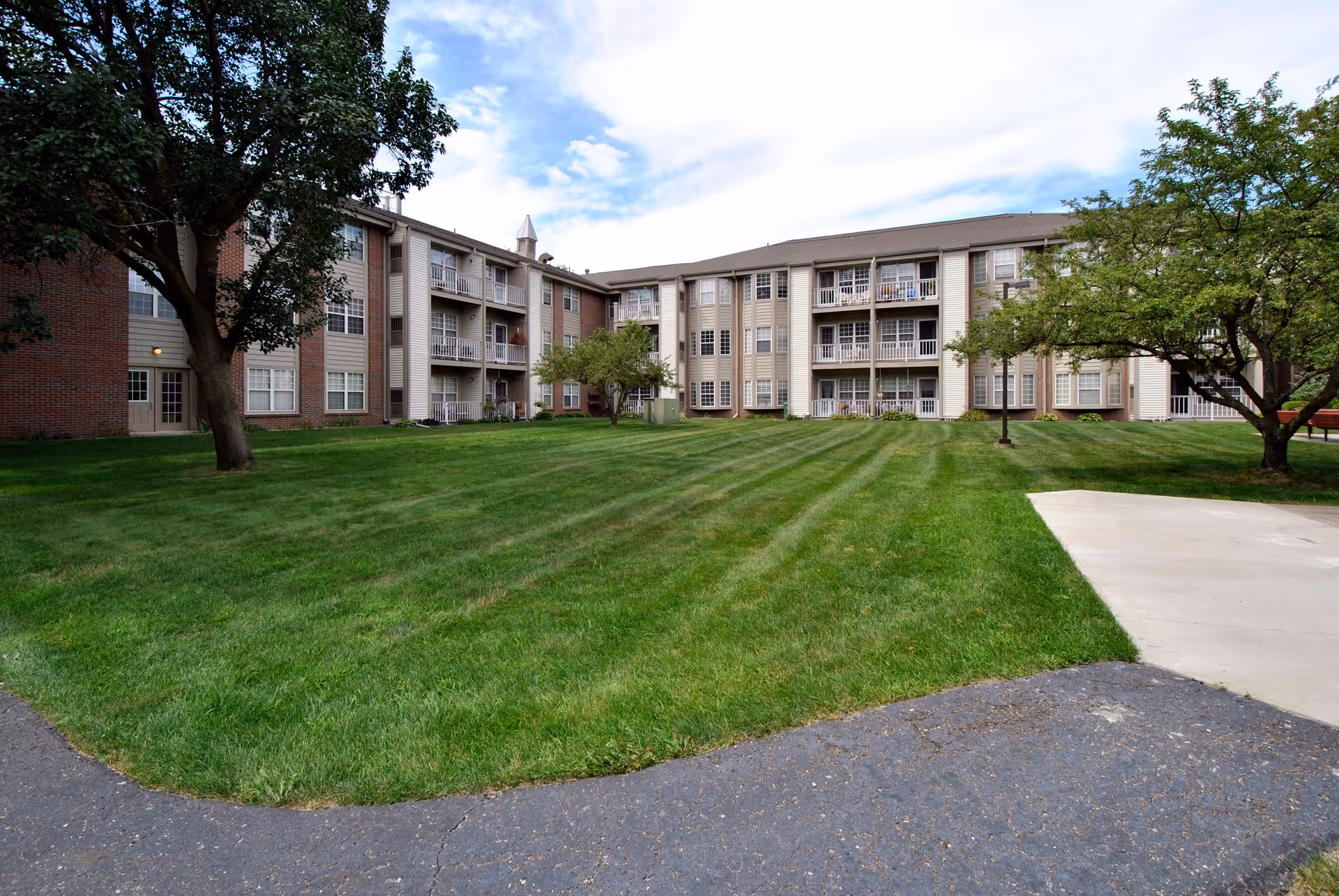 A large, well-maintained grassy courtyard area surrounded by a three-story senior living facility building with balconies and windows. There are several trees and a paved walkway around the grass under a partly cloudy sky.
