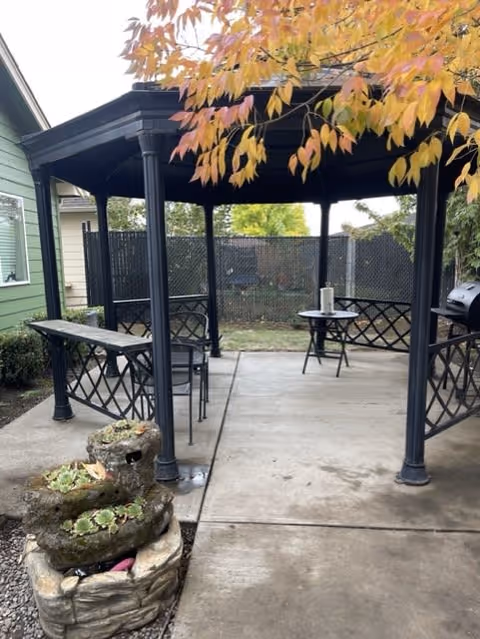 Outdoor covered patio area with a black metal gazebo structure, a small round table with a paper towel roll on it, and a stone planter with succulents. The area is surrounded by a fence and there are trees with autumn-colored leaves overhead.