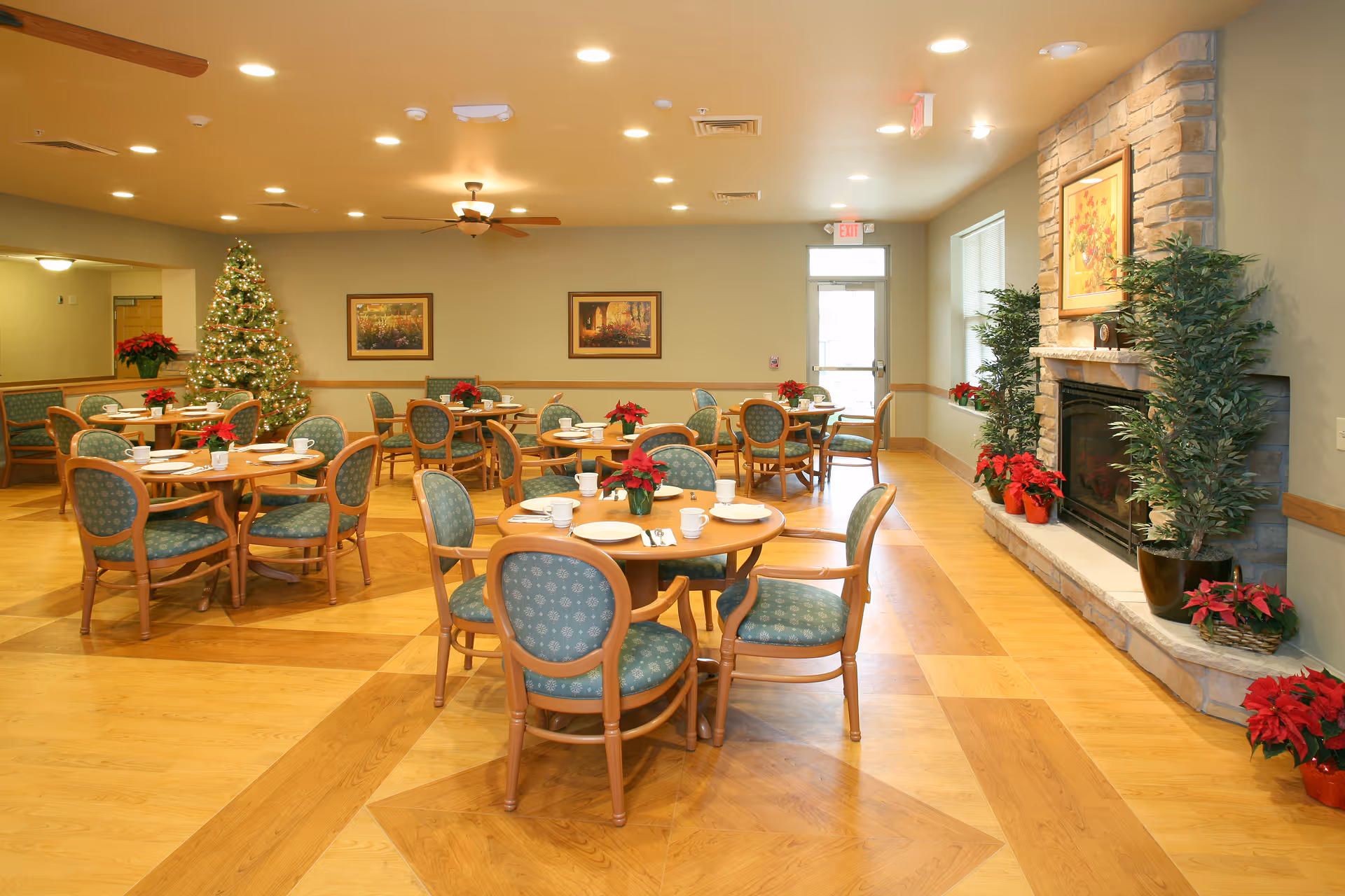 A bright dining room with several round wooden tables and green cushioned chairs arranged neatly. Each table is set with white plates, cups, and silverware, and decorated with red poinsettia plants. There is a decorated Christmas tree in the corner, framed paintings on the walls, and a stone fireplace adorned with plants and more poinsettias. The room has wooden flooring and ceiling fans with lights.