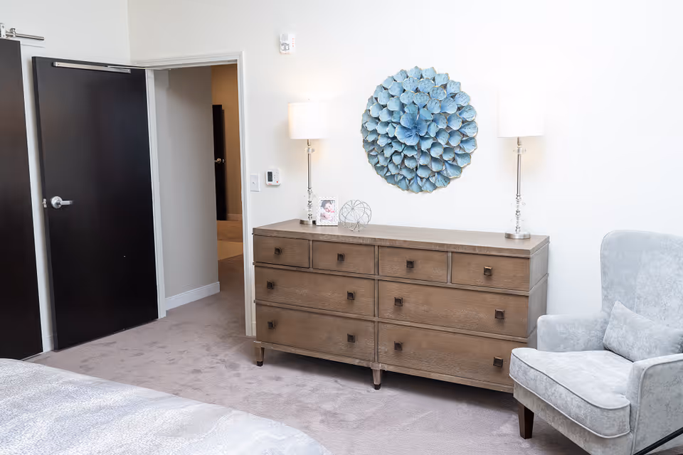 Bright bedroom interior with a wooden dresser topped by lamps and decorative wall art, a gray armchair, and an open doorway.