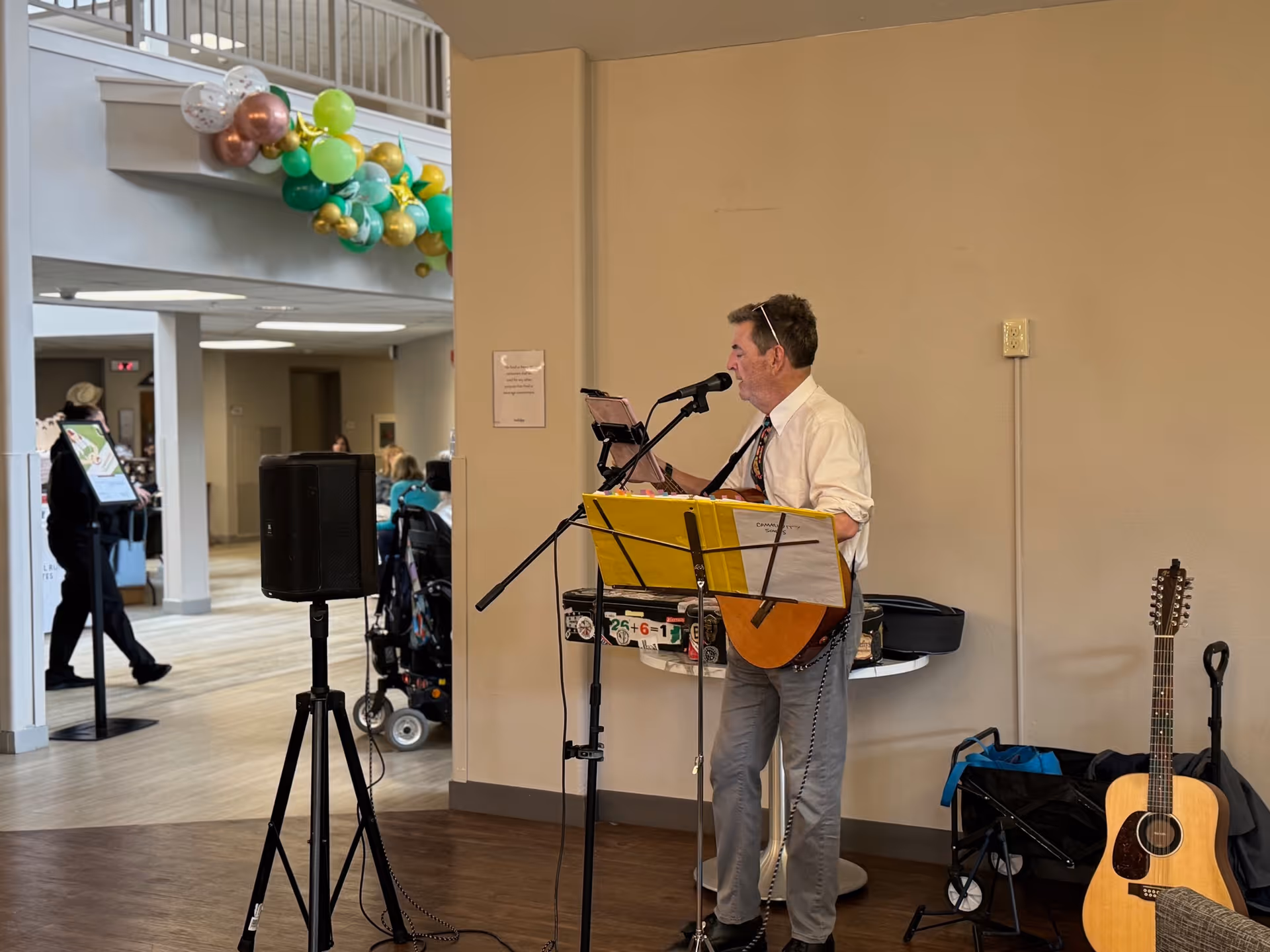 A man wearing a white shirt and tie is playing a guitar and singing into a microphone in an indoor common area. There is a music stand with sheet music in front of him, a speaker on a tripod stand nearby, and another guitar resting against a chair. In the background, there are people sitting and walking in a spacious, well-lit area decorated with green, gold, and white balloons.