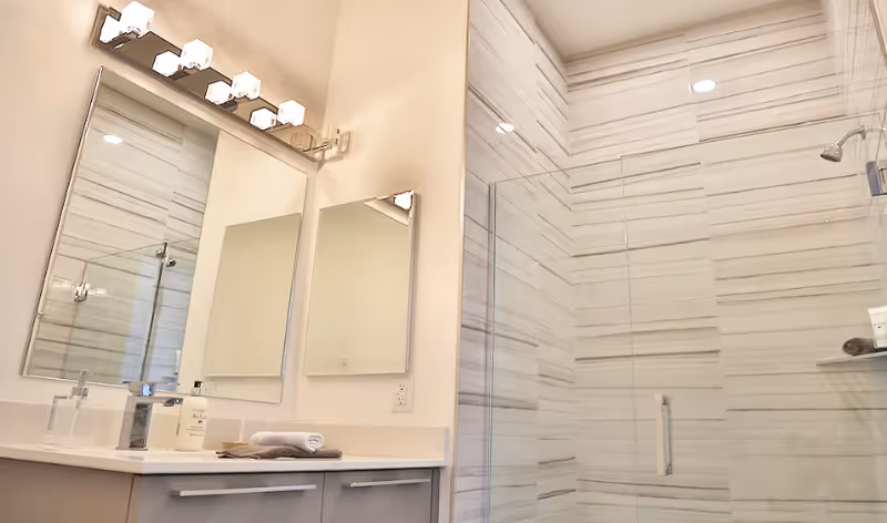 Modern bathroom with a large mirror above a white countertop with a sink, soap dispenser, and folded towels. Next to the sink is a glass-enclosed shower with light-colored horizontal striped tiles and a chrome showerhead.