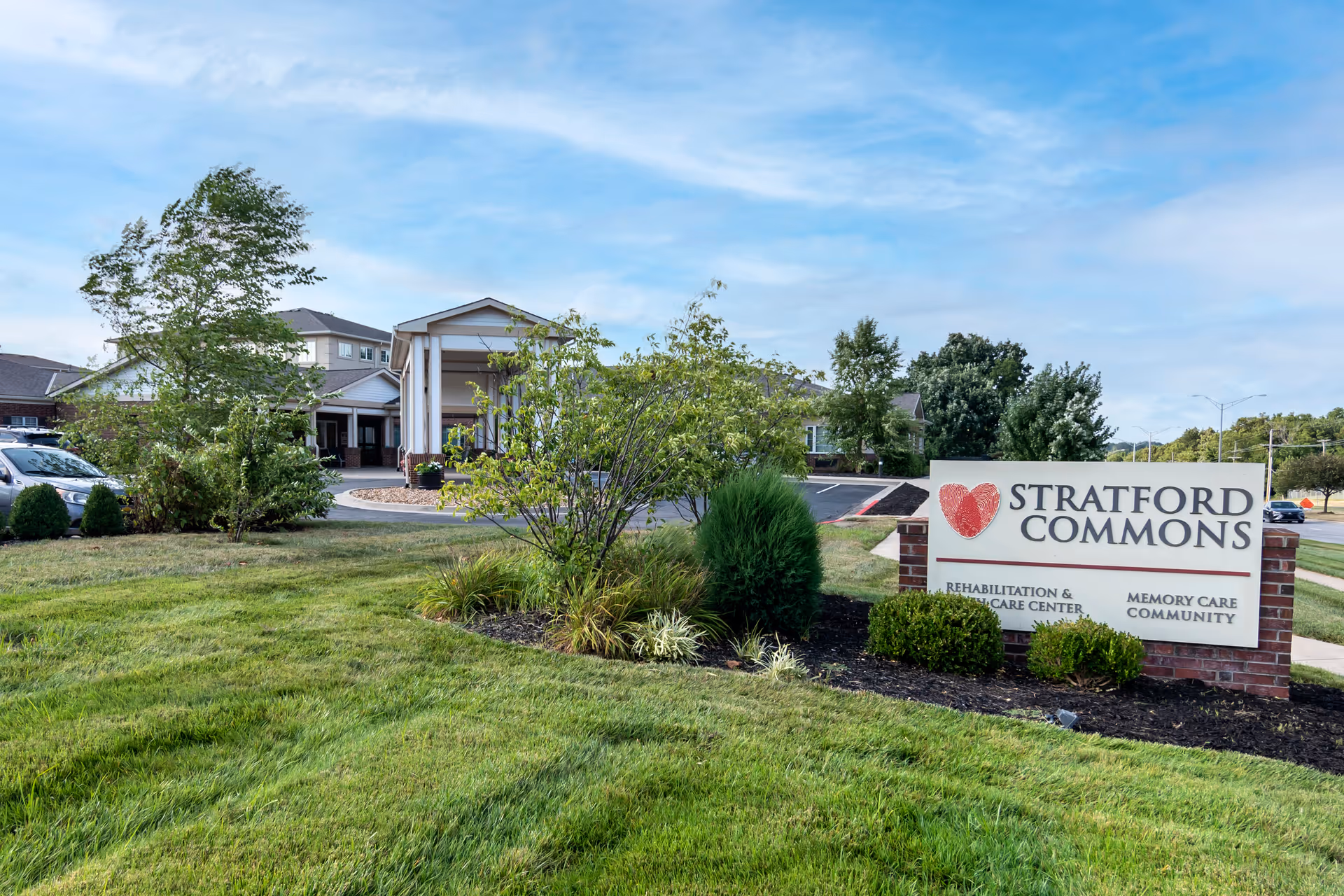 Exterior view of Stratford Commons Rehabilitation & Health Care facility with a landscaped lawn, trees, and a sign displaying the facility name and services including rehabilitation, health care, and memory care community.