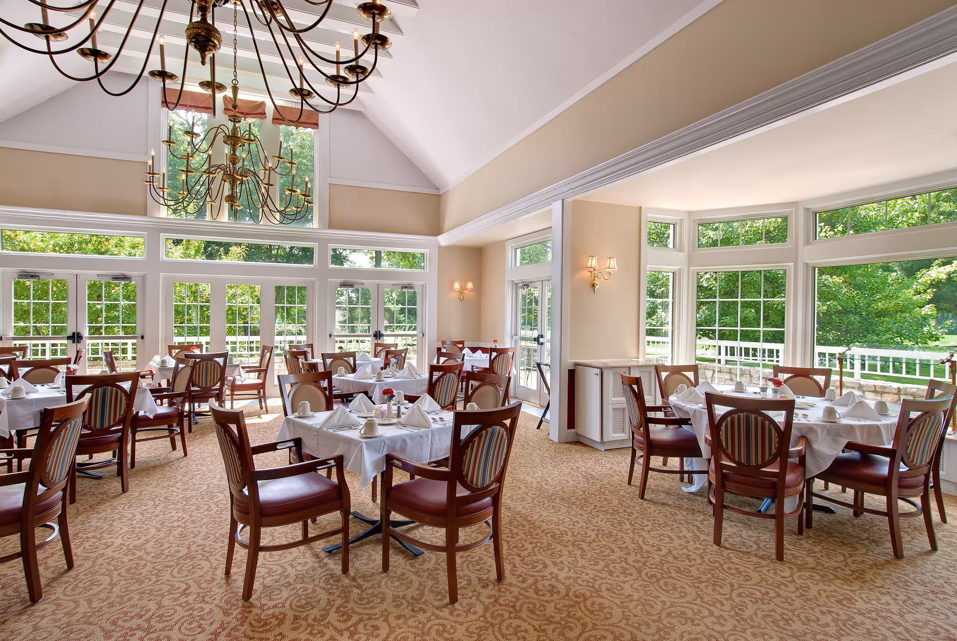 Bright and spacious dining room with multiple tables covered in white tablecloths, each set with napkins, plates, and small flower vases. Large windows surround the room, allowing natural light to fill the space and offering views of green trees outside. A large chandelier hangs from the high ceiling, and the room has beige walls and patterned carpet flooring.