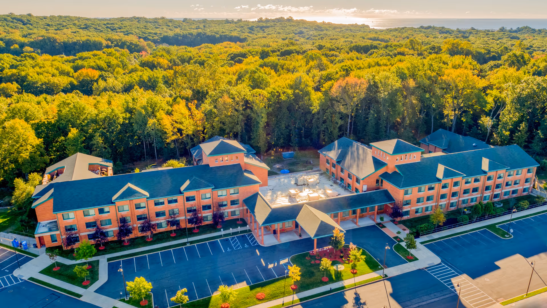 Aerial view of The Brielle senior living facility surrounded by dense green trees. The building is large with multiple wings and a dark roof, featuring a covered entrance and adjacent parking lots with marked spaces. The scene is bathed in warm sunlight with a clear sky.