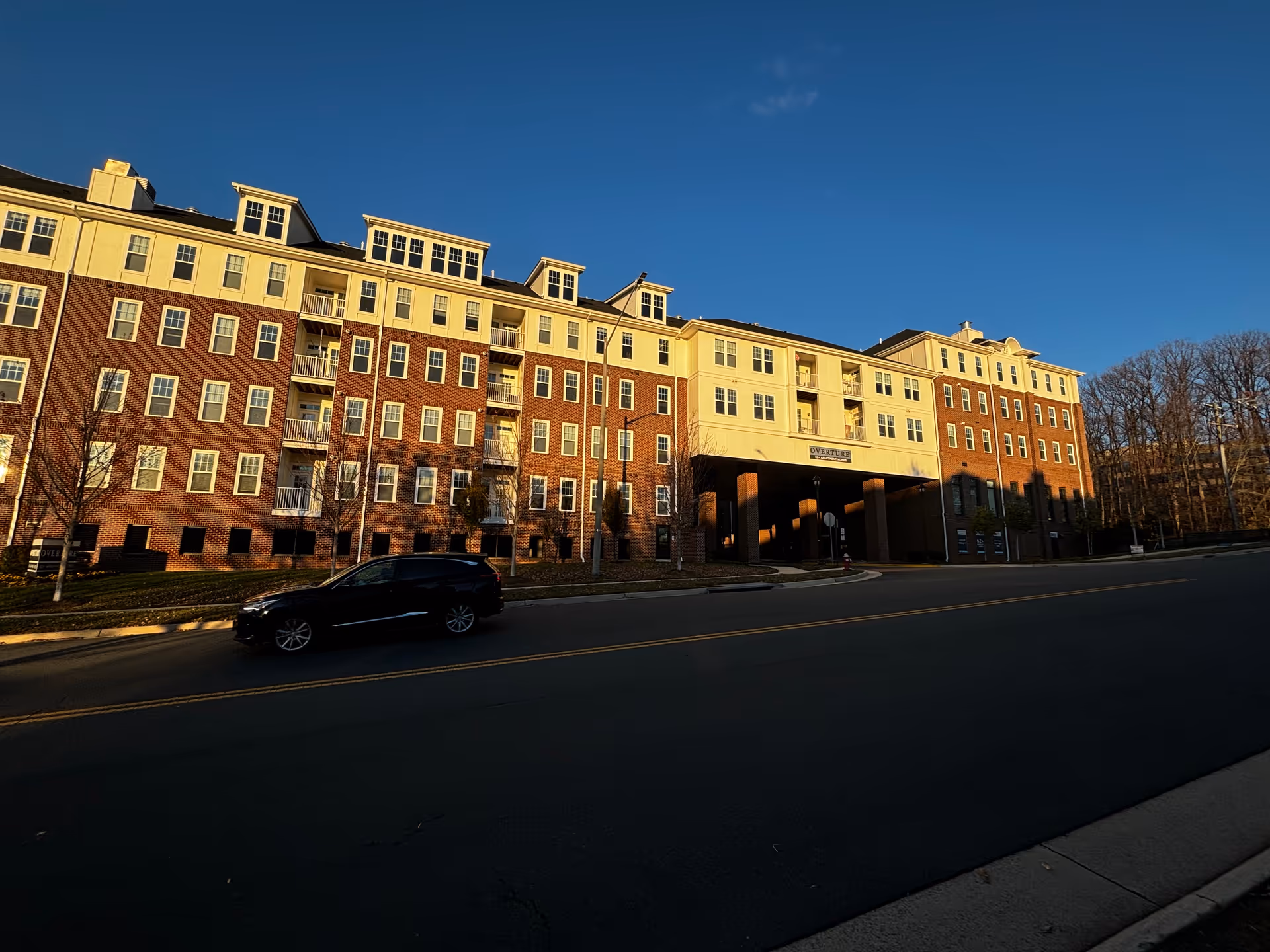 Exterior view of a multi-story senior living facility building named Overture Fair Ridge, with a brick and cream-colored facade, balconies, and a driveway in front. A black car is parked on the street, and trees without leaves are visible on the right side under a clear blue sky.