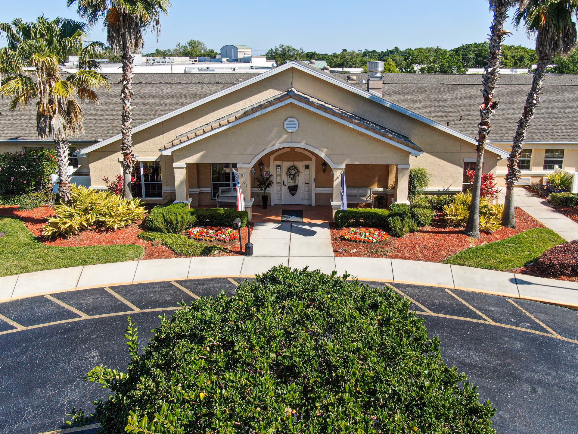 Front exterior view of a single-story building with a peaked roof, a covered entrance with two white benches, and landscaped flower beds with palm trees and shrubs. A circular driveway is visible in front of the entrance.