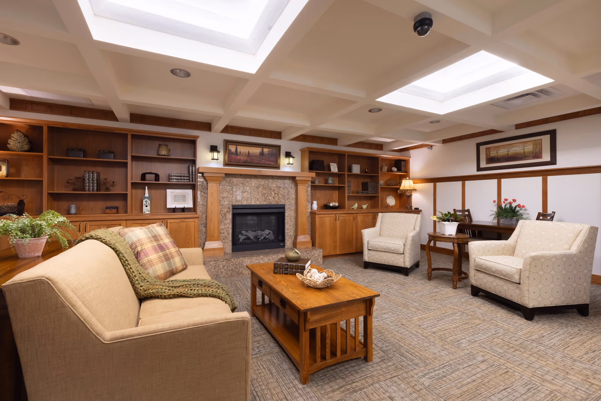 Cozy common room with sofas, armchairs, a coffee table, built-in wooden bookshelves and a fireplace under skylights.