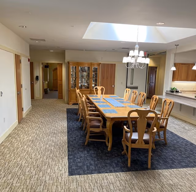 Long wooden dining table with chairs and placemats under a skylight and chandelier in a communal dining area.