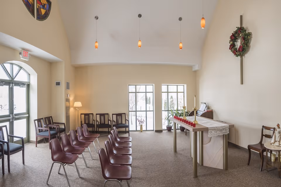 Interior of a small chapel or prayer room with rows of chairs facing an altar decorated with a lace cloth, candles, and a wreath hanging on the wall above a cross. There are large windows letting in natural light, a lamp in the corner, and stained glass decorations near the ceiling.