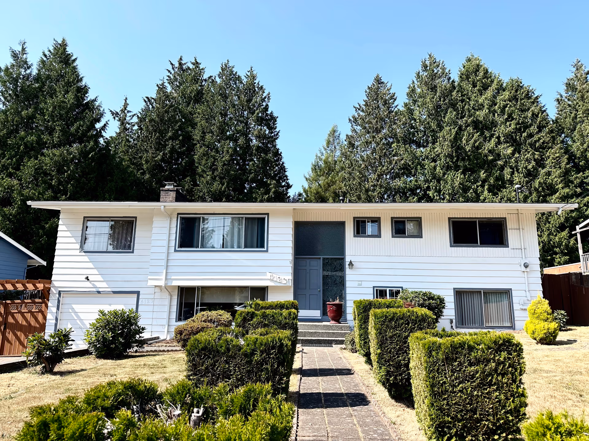 Front exterior view of a white two-story house with a gray door, multiple windows, and a well-maintained garden with trimmed bushes and a paved walkway leading to the entrance. Tall evergreen trees are visible in the background under a clear blue sky.